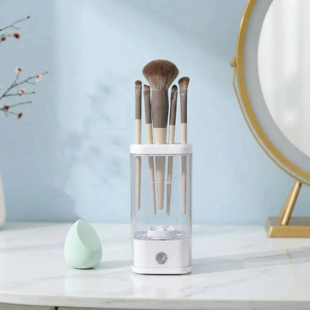 CleanLuxe Automatic Makeup Brush Cleaner in white with beige handled brushes, a clear container, and a control button on a marble surface